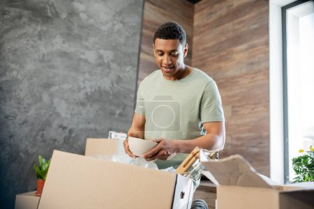 Photo for Joyful african american man holding plate while unpacking cardboard boxes in new house - Royalty Free Image