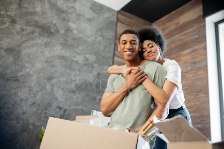 lächelnde Afroamerikanerin umarmt Freund, während sie in der Nähe von Paketen in neuem Haus steht