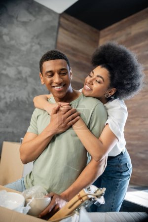 alegre africana americana mujer abrazando novio cerca de caja de cartón en nueva casa