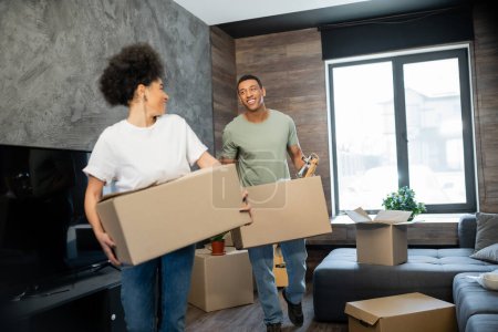 alegre pareja afroamericana sosteniendo paquetes de cartón durante la reubicación en nueva casa
