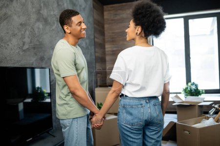 pareja afroamericana llena de alegría cogida de la mano mientras está de pie cerca de cajas de cartón en casa nueva