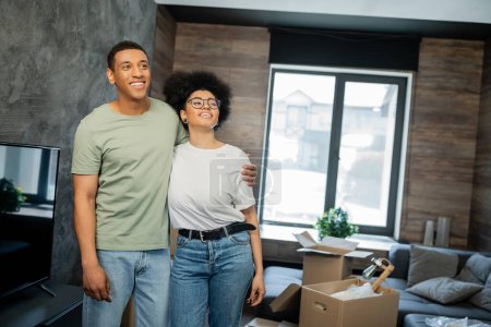 Téléchargez les photos : Couple afro-américain rêveur et souriant étreignant près de boîtes de caron dans une nouvelle maison - en image libre de droit