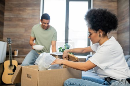 Photo for African american woman unpacking carton box near blurred boyfriend during relocation in new house - Royalty Free Image