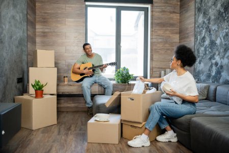 lächelnder afrikanisch-amerikanischer Mann spielt Akustikgitarre in der Nähe von Freundin und Kartons in neuem Haus