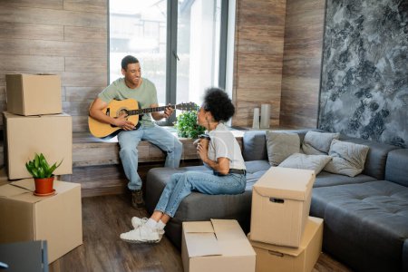 fröhliches afrikanisch-amerikanisches Paar mit Kaffee spielt Akustikgitarre in der Nähe von Boxen im neuen Haus