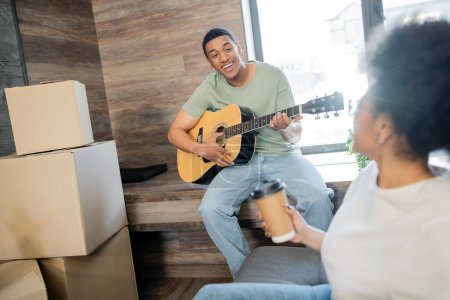 hombre afroamericano feliz tocando la guitarra acústica cerca de novia borrosa con café en casa nueva