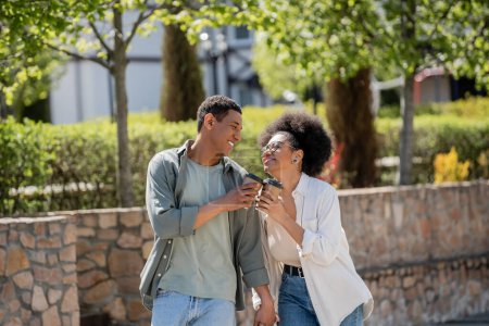 Téléchargez les photos : Joyeux couple afro-américain tenant la main et café pour aller tout en marchant dans la rue urbaine - en image libre de droit