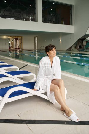 Frau mittleren Alters mit kurzen Haaren sitzt auf einer Liege, weißer Bademantel, Wellness-Center, innen, Schwimmbad