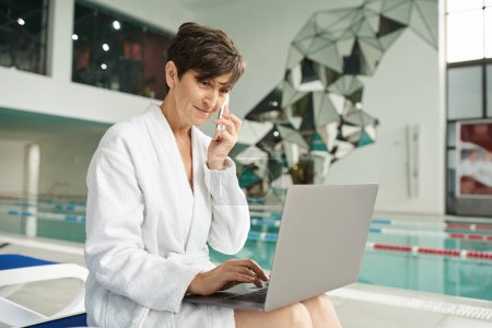 freelancer, middle aged woman talking on smartphone, using laptop, sitting on lounger, spa center