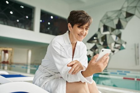 spa center, happy middle aged woman using smartphone, sitting on lounger, white robe, swimming pool