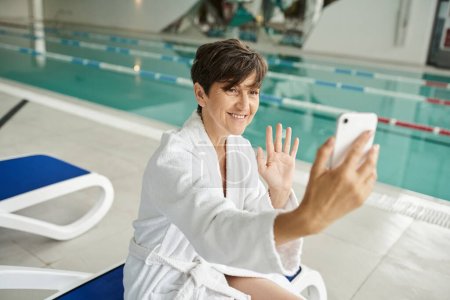 positive middle aged woman having video call on smartphone, lounger, swimming pool, spa center