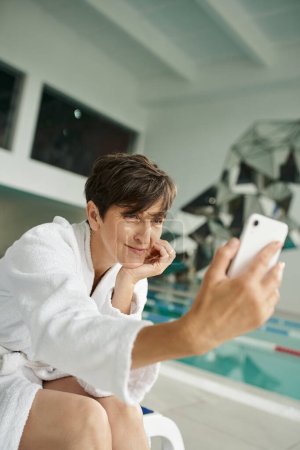 Foto de Mujer positiva de mediana edad en bata blanca tomando selfie, teléfono inteligente, centro de spa, piscina - Imagen libre de derechos