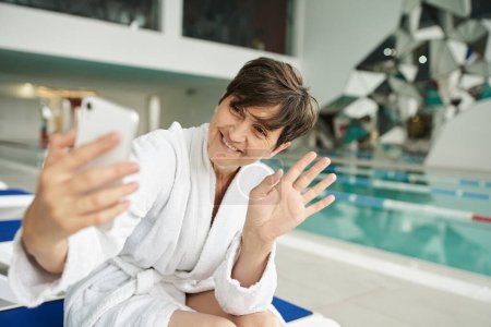 Téléchargez les photos : Femme d'âge moyen positif ayant un chat vidéo sur smartphone, main vague, piscine, centre de spa - en image libre de droit