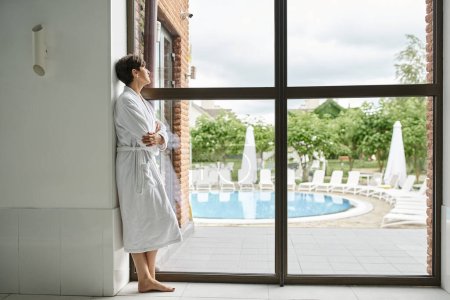 middle aged woman with short hair standing in white robe near panoramic window in spa center, pool
