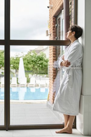 reife Frau mit kurzen Haaren in weißer Robe in der Nähe von Panoramafenster im Wellness-Center, Blick auf den Pool