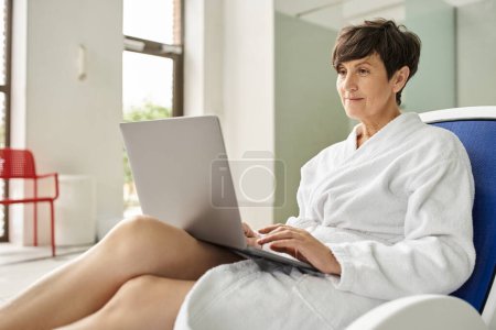 Frau mittleren Alters in weißem Gewand mit Laptop, auf Liegen im Wellness-Center sitzend, Fernarbeit, drinnen