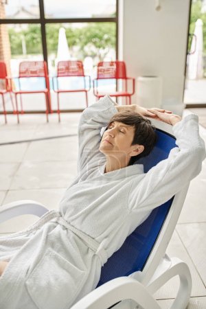 middle aged woman with short hair relaxing on lounger, white robe, spa center, closed eyes