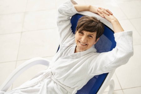 glückliche Frau mittleren Alters mit kurzen Haaren entspannt auf Liege, weißer Robe, Wellness-Center, Blick in die Kamera