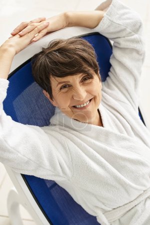 Photo for Happy mature woman with short hair relaxing on lounger, white robe, spa center, look at camera - Royalty Free Image