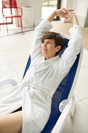 joyful middle aged woman with short hair stretching on lounger, white robe, spa center, smile