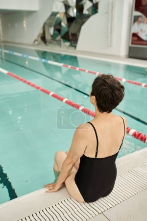 back view of tattooed woman with short hair sitting in swimsuit, swimming pool, poolside, spa center