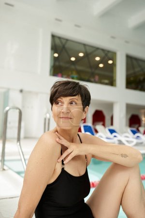 portrait of tattooed middle aged woman in swimsuit sitting near swimming pool, spa center, look away