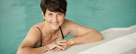 cheerful and mature woman with tattoo swimming in pool, indoors, spa center, wellness, banner