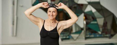 cheerful mature swimmer in swimsuit adjusting goggles, woman in swim cap, wellness center, banner
