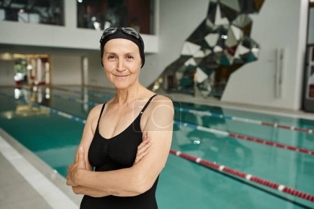 cheerful middle aged woman in swim cap and goggles, swimming pool in wellness center, folded arms