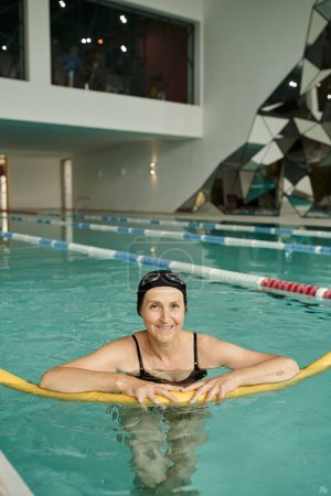 happy middle aged woman in swim cap and goggles swimming with pool noodle, healthy lifestyle