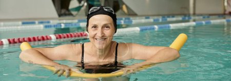 happy mature woman in swim cap and goggles swimming with pool noodle, healthy lifestyle, banner