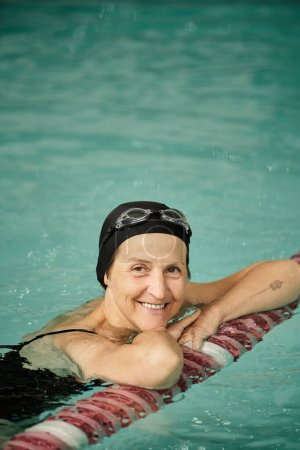 positive Frau mittleren Alters schwimmt im Pool, Badekappe und -brille, blickt in die Kamera, Sport, Tätowierung