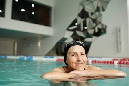 happy middle aged woman swimming with floating board in pool, swim cap and goggles, smile