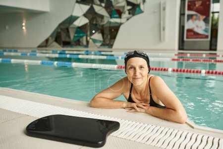happy woman leaning on poolside near floating board, swim cap and goggles, smile, middle aged