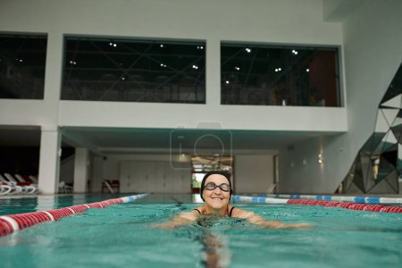 cheerful middle aged woman in swim cap and goggles swimming in pool, recreation center, lifestyle