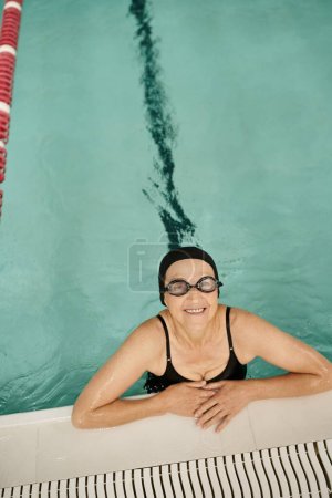 top view of happy middle aged woman in swim cap and goggles, blue water in pool, recreation center