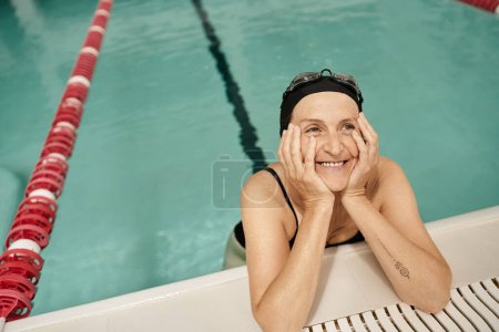 middle aged woman in swim cap and goggles looking away, smiling, water in pool, recreation center