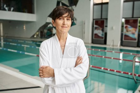 Foto de Mujer de mediana edad con el pelo corto de pie con los brazos cruzados cerca de la piscina, bata blanca, centro de spa - Imagen libre de derechos
