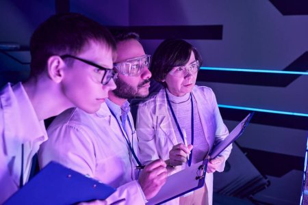 Foto de Análisis futurista: Tres científicos profundizan en el estudio de dispositivos en el Neon-Lit Science Center. - Imagen libre de derechos