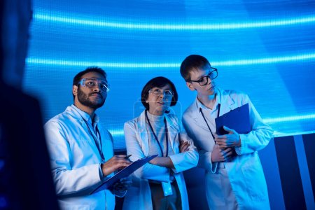 Zukunftsorientierte multiethnische Wissenschaftler mit Klemmbrettern arbeiten in neonbeleuchtetem Innovationszentrum zusammen