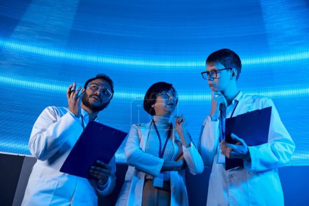 futuristische Lösungen, multiethnische Wissenschaftler mit Klemmbrettern im neonbeleuchteten Entdeckungszentrum