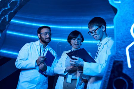 Junge Praktikantin blickt in neonbeleuchtetem Entdeckungszentrum auf Klemmbrett neben multiethnischen Kollegen