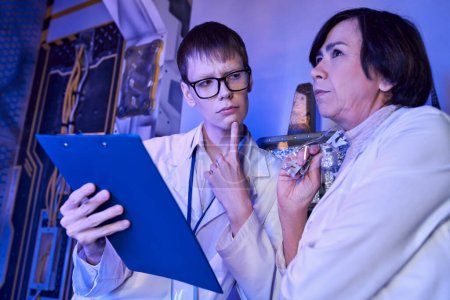 Nachdenkliche Praktikantin mit Klemmbrett blickt auf Wissenschaftlerin mittleren Alters in futuristischem Wissenschaftszentrum