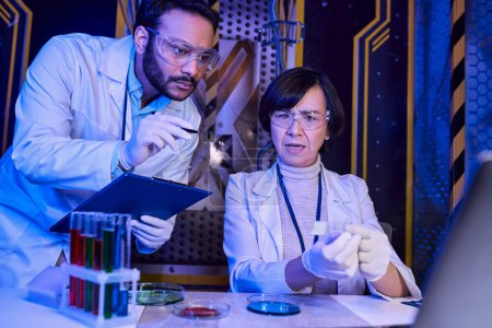 Indischer Wissenschaftler zeigt mit Pfanne auf Proben in der Nähe eines Kollegen in futuristischem wissenschaftlichen Labor