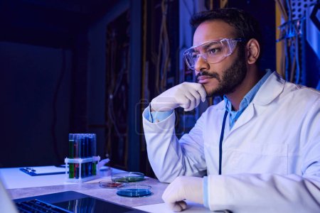 Indischer Wissenschaftler mit Brille arbeitet in futuristischem Labor an Laptop in der Nähe von Reagenzgläsern und Petrischalen