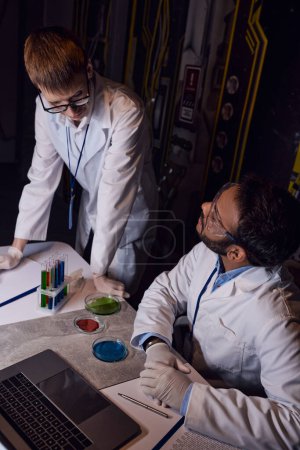 científicos multiétnicos que trabajan cerca de tubos de ensayo, placas de Petri y portátil en laboratorio innovador