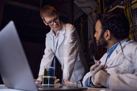 Multiethnische Wissenschaftler sprechen in futuristischem Labor in der Nähe von Laptop und Reagenzgläsern mit Proben außerirdischen Lebens