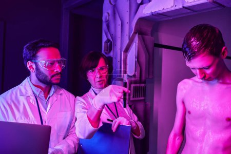 Multiethnische Wissenschaftler betrachten Reagenzglas mit Blutprobe in der Nähe von humanoidem Alien im Science Center