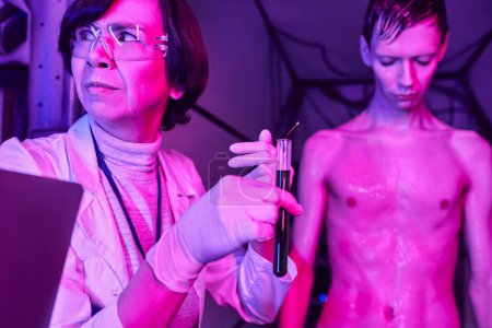 woman scientist in goggles holding test tube with blood sample near humanoid alien in innovative lab