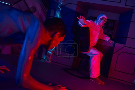 Téléchargez les photos : Humanoïde extraterrestre rampant près du scientifique en combinaison hazmat montrant geste d'arrêt en laboratoire - en image libre de droit